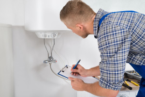 Male Plumber Writing On Clipboard In Front Of Electric Boiler In Kitchen Room
